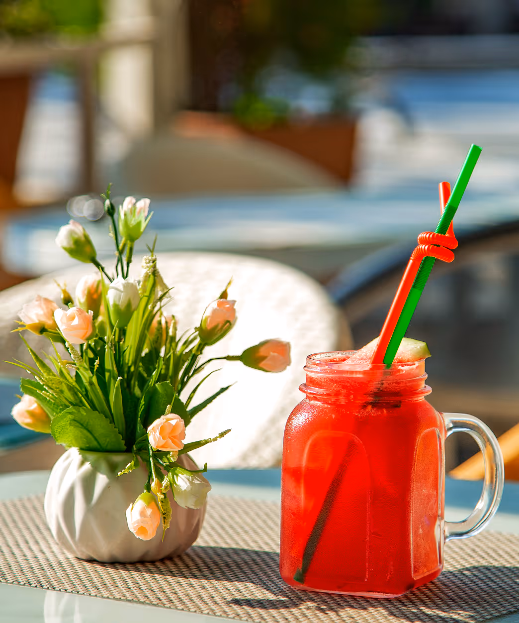 Red iced drink with green and red straws in a glass jar next to a vase of pale pink flowers on a round table.